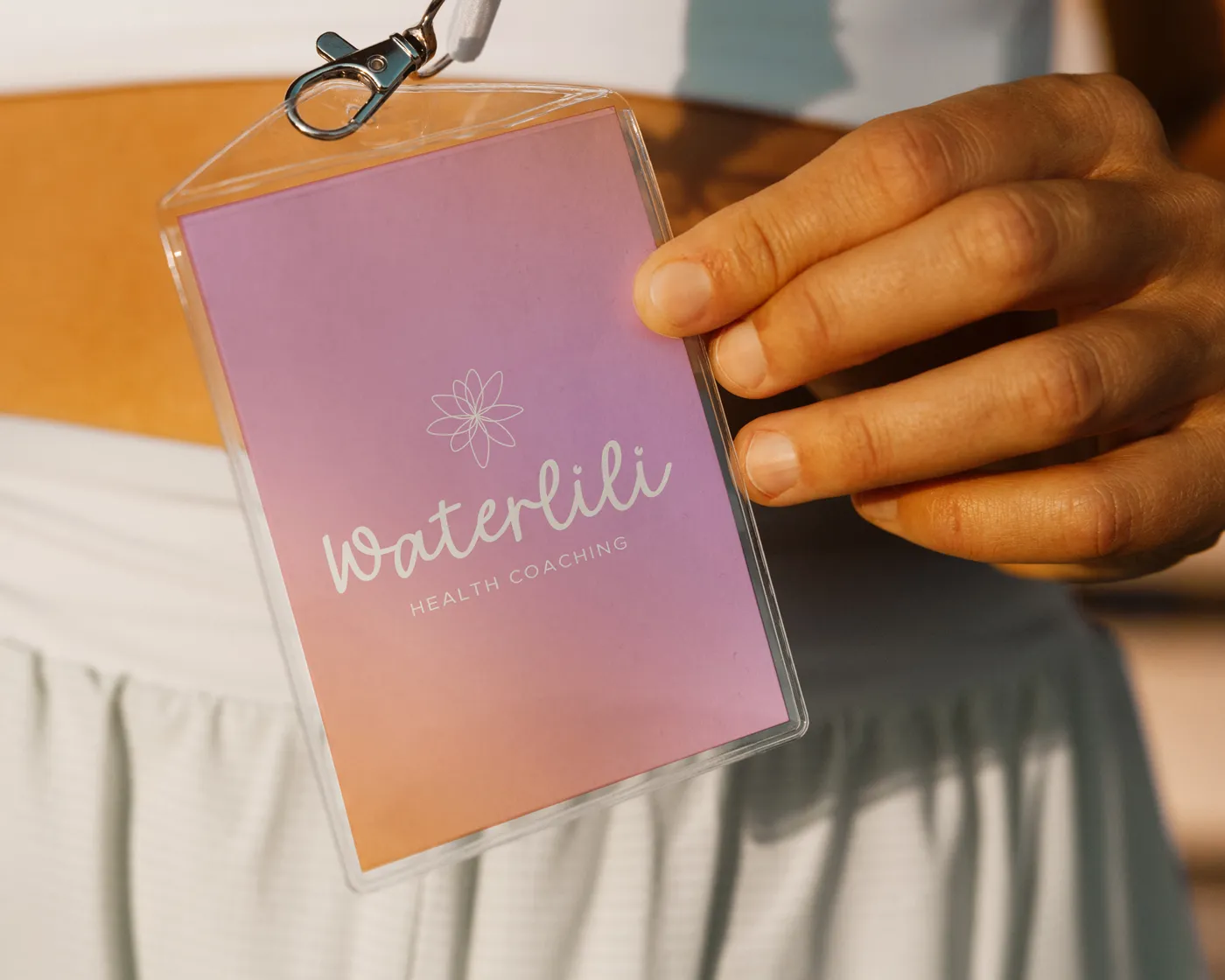 A person in athletic wear holds a pastel gradient badge with the Waterlili Health Coaching logo and name. The background is a soft ombre blend of pink, orange, and purple with a delicate flower icon above the text.
