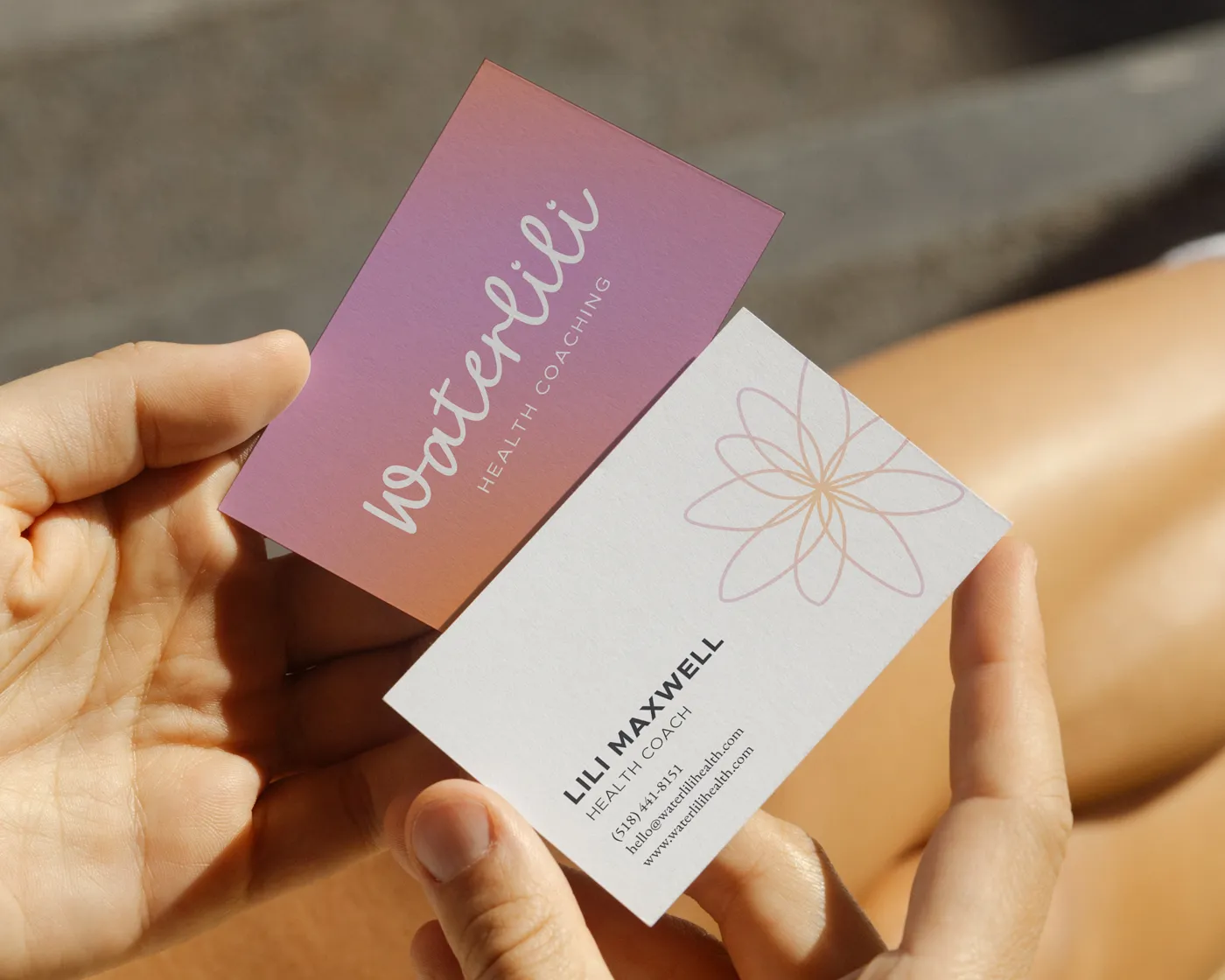 Hands holding two business cards for Waterlili Health Coaching. One card has a minimalist logo and gradient background, while the other displays contact details for Lili Maxwell, Health Coach, with a soft floral design in the top right corner.