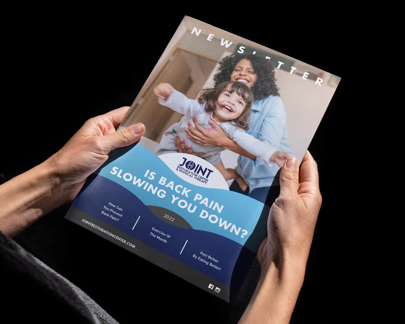 Hands holding a newsletter for Joint Restoration Center & Physical Therapy. The cover shows a joyful woman playing with a young girl in a home setting, with large text reading “Is Back Pain Slowing You Down?” A blue curved graphic highlights sections inside: “How Can You Prevent Back Pain?”, “Exercise of the Month,” and “Feel Better by Eating Better!”